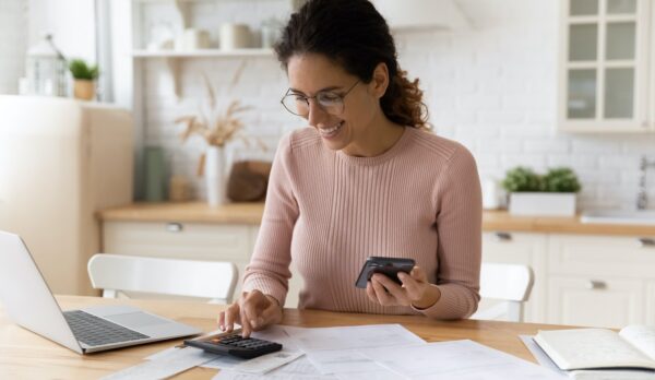 Young latina female work with financial papers at home count on calculator before paying taxes receipts online by phone. Millennial woman planning budget glad to find chance for economy saving money